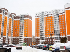 многоэтажные современные дома новостройки с ярко-желтыми стенами по центру, коричневыми внизу дома и на верхних этажах с детской игровой площадкой и машинами на парковочных местах во дворе в зимнее время