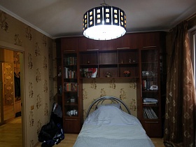 мебельная стенка с многочисленными полками над кроватью с металлической спинкой в спальне с черным абажуром на белом потолке актерской трехкомнатной квартиры