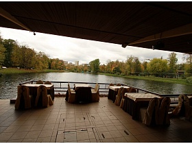  бежево коричневые чехлы на стульях вокруг уютных квадратных столиков с длинной коричневой скатертью и короткой бежевой сверху на плиточном полу под крышей открытого кафе на озере