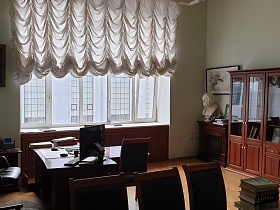 общий вид строгого кабинета КГБ СССР с рабочим столом, столом для заседаний, длинным шкафом для книг, белым бюстом на деревянном камине в углу о окна с белым ламбрекеном