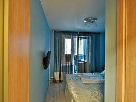 темно серые гардины на окне с балконной дверью и телевизор на голубой стене спальной комнаты молодежной квартиры высотного здания в Новострое