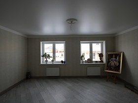 картина в рамке на мольберте в углу светлой комнаты с зелеными комнатными цветами на подоконниках окон без штор