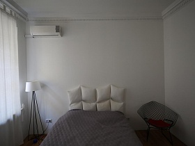 белый торшер на черных высоких ножках, белая большая кровать с серым покрывалом и необычное сетчатое кресло у белой стены светлой спальни скандинавской квартиры