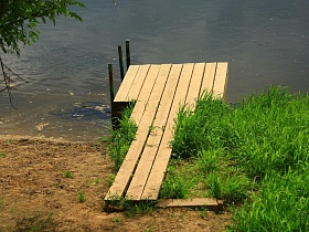 деревянный настил мостика на металлических опорах на песчанном берегу реки среди зеленой травы