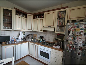 белая мебельная стенка с многочисленными шкафами, встроенной газовой плитой, телевизором на коричневой столешнице, серебристый холодильник у белой стены кухни сталинской квартиры 90 годов