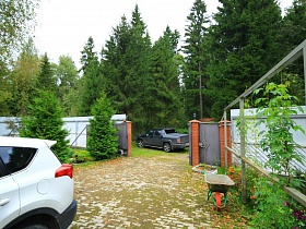 машины во дворе с брусчаткой и за забором семейного двухэтажного дома среди густого хвойного леса