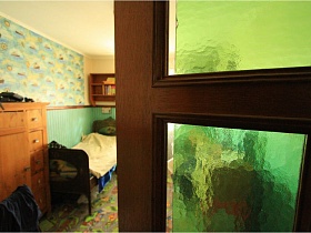 коричневый шкаф, кровать со спинками, навесная полка на стене у окна детской сквозь приоткрытую дверь со стеклянными вставками