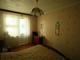 песочный светильник над большой кроватью и плоский телевизор на коричневом комоде в углу спальной комнаты трешки