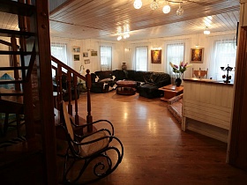 плетенное кресло-качалка у деревянной лестницы с резными перилами в просторной светлой гостиной, освещенной множеством окон на классической семейной даче
