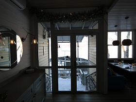 бра со стеклянными плафонами у круглого зеркала над белым шкафчиком у стены рядом с входными дверьми со стеклянными вставками в здание белого ресторана на берегу озера