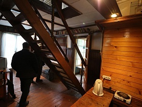 деревянная лестница с перилами на второй этаж в зонированной комнате небольшого домика отшельника среди новостроек