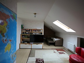 синяя карта мира на белой стене комнаты отдыха со светлым ковром у диванов и мебельной стенки с телевизором в мансарде