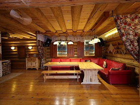 общий вид уютной зоны отдыха за цветными шторами с красным угловым диваном с подушками вокруг деревянных столов  у стены с полотенцами и окон со ставнями