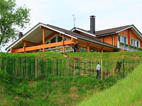 красивый двухэтажный деревянный дом с открытой террасой за решетчатым металлическим забором на крутом зеленом берегу реки с изгибом и островками