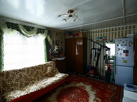 сортивные снаряды у стены с зеркалом, серым холодильником и шкафами в углу гостиной деревянной избы в деревне