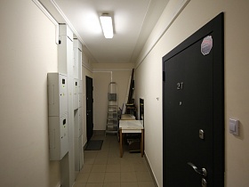 лестница-стремянка, стол, коробки на полу светлого длинного коридора на первом этаже жилого дома с электро щитовыми коробками на стене и входными дверьми квартир