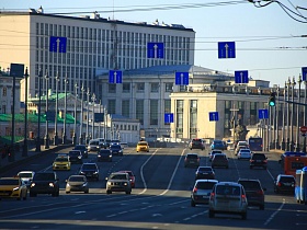 восьмиполосная оживленная трасса на большом каменном мосту с рядами высоких столбов с фонарями вдоль дороги с общественным городским транспортом и легковыми машинами у Кремля