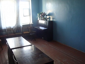 коричневые столы в центре на деревянном полу, черное пианино у стены, окрашенной в голубой цвет, стулья со спинкой у высоких  окон музыкальной комнаты старого клуба времен СССР
