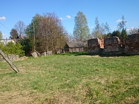 стены разрушенного кирпичного здания на открытой площадке у покосившегося деревянного забора вокруг жилого дома в старой деревне Троица