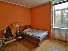 бежевое кресло, торшер, деревянная кровать в углу спальни с ораньжевыми стенами в трехкомнатной квартире №16