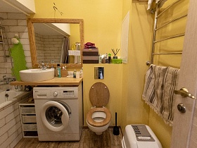 белая ванна зашитая кирпичом у кирпичной стены,стиральная машинка под деревянной столешницей с белой раковиной и зеркалом в деревянной рамке на стене, полотенцесушитель в ванной комнате с желтыми стенами современной квартиры в сталинском доме