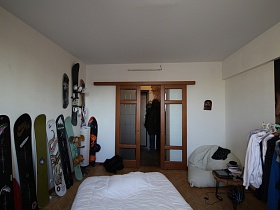 кровать под белым стеганным одеялом в центре комнаты, белое кресло в углу комнаты после ремонта с деревянными раздвижными дверьми со стеклянными вставками трехкомнатной квартиры