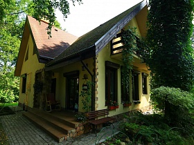 плитка на земле вокруг желтого двухэтажного домика с открытым балконом на зеленом дачном участке