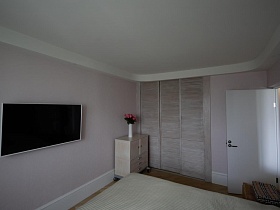 плоский белый телевизор на стене напротив большой кровати,розы в вазочке на тумбе у встроенного шкафа-купе в розовой спальне с белым потолком стильной трехкомнатной квартиры