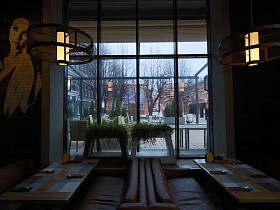 круглые сетчатые абажуры подвесных светильников над индивидуальными столиками с мягкими креслами у большого окна с комнатными цветами на оригинальных стойках крафтового ресторана