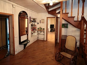 зеркало в рамке с полочкой у двери, встроенный шкаф с полками, кованная этажерка с фотографиями в углу гостиной под деревянной витой лестницей с перилами и рядом стоящим плетенным креслом в гостиной семейной дачи