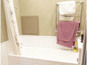 полотенцесушитель с розовым полотенцем и белая шторка над зашитой белой ванной на белой с черным плиткой на полу выанной комнаты в двушке жилого дома