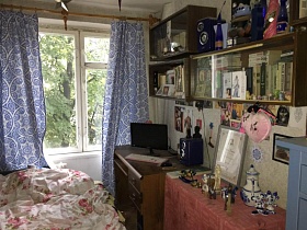 многочисленные сувениры, диплом в рамке на столе, ноутбук и колонки на компьютерном столе у светлой стены с деревянными книжными полками спальной комнаты двухкомнатной квартиры