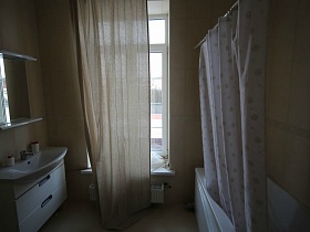 зашторенное окно ванной комнаты с белой ванной за шторкой , зеркалом с полкой над белым шкафом кирпичного двухэтажного дома