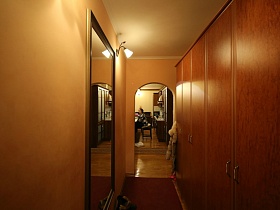 высокое прямоугольное зеркало в рамке, бра на персиковой стене прихожей с коричневым шкафом, арочным дверным проемом на кухню модной дизайнерской квартиры