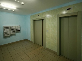 серые почтовые ящики на голубой стене просторного холла с двумя лифтовыми кабинами и квадратной плиткой на полу современного многоэтажного дома в новострое