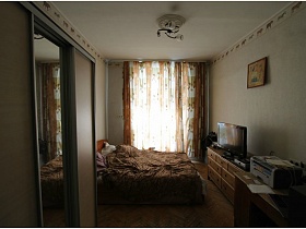 картина над комодом с телевизором, большая кровать с коричневым покрывалом и коричневые цветные шторы на окне белой спальной комнаты типичной двушки