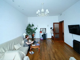 высокие комнатные цветы в углу, белый столик и напольная вешалка для одежды за открытой дверью в гостиную с черным телевизором на стене белой гостиной современной евро квартиры