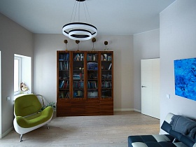 книжный шкаф со стеклянными дверцами, зеленое мягкое кресло и синяя абстакция на стене в гостиной