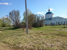 белое здание церкви с крестами на синих куполах за деревянным забором с широкими воротами и белыми колонами в старой деревне Троица