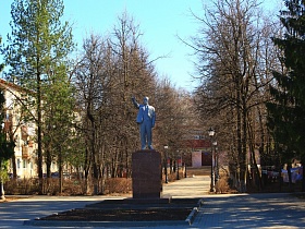 серый памятник Ленина на высоком постаменте на площади, выложенной плиткой и ухоженными клумбами с зелеными высокими елями вокруг