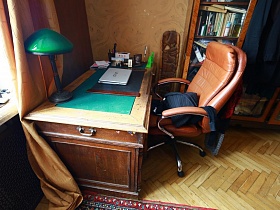 ноутбук и настольная лампа с зеленым абажуром на старинном письменном столе и коричневое компьютерное кресло  в углу спальни трехкомнатной советской квартиры