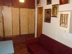 голубой квадратный стол напротив дивана с вишневым покрывалом, большие портреты и картины в рамках на белой стене, высокий шкаф для одежды у открытой двери светлой комнаты квартиры бабушки эпохи СССР