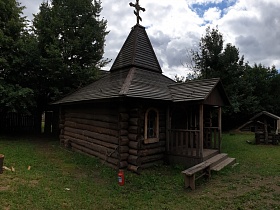 небольшая деревянная часовня с крестом наверху, открытым деревянным крыльцом во дворе среди сосен за деревянным забором в небольшой деревне