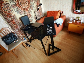 ноутбук на черном стеклянном столике и колонки на подставках, ораньжевое кресло, сумка для ноутбука на стуле со спинкой у стены с цветным ковром в спальне квартиры сталинки