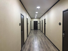 бежево коричневый линолеум под дерево на полу длинного светлого коридора с круглыми светильниками на белом потолке, с коричневыми  номерками у входных дверей в номера гостиницы
