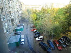 парковачные места с машинами вдоль зеленого участка с высокими деревьями в придомовой территории многоэтажки