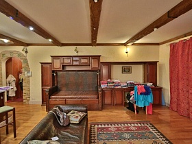 коричневая мебельная стенка со встроенным диваном , цветной ковер у мягкого дивана зонированной гостиной в просторной кухне с декоративным камнем вокруг арочного проема