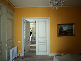 электрический радитор у белого встроенного шкафа для одежды, картина с пейзажом на желтой стене с открытой дверью спальной комнаты современной дизайнерской двухкомнатной квартиры