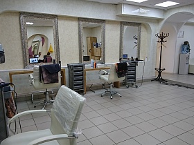 ряд оборудованных рабочих мест, сушилка для волос, напольная вешалка для одежды на полу со светлой квадратной плиткой просторного современного салона парикмахерской 2