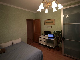 шкаф для одежды с рифленным стеклом на дверцах, комнатный цветок, белый телевизор и ноутбук на белом столе у стены с картинами за дверью сиреневой спальни простой квартиры молодоженов